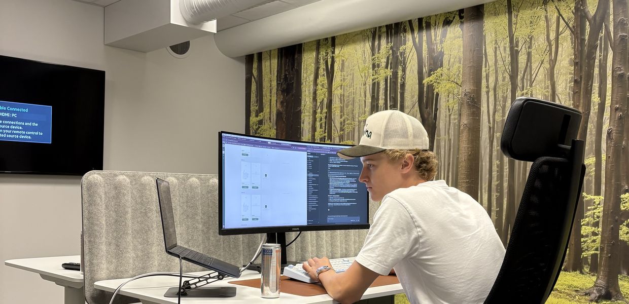 En av grundarna vid skrivbordet i studion, mitt i ett bygge — kod och skisser uppe på dubbla skärmar